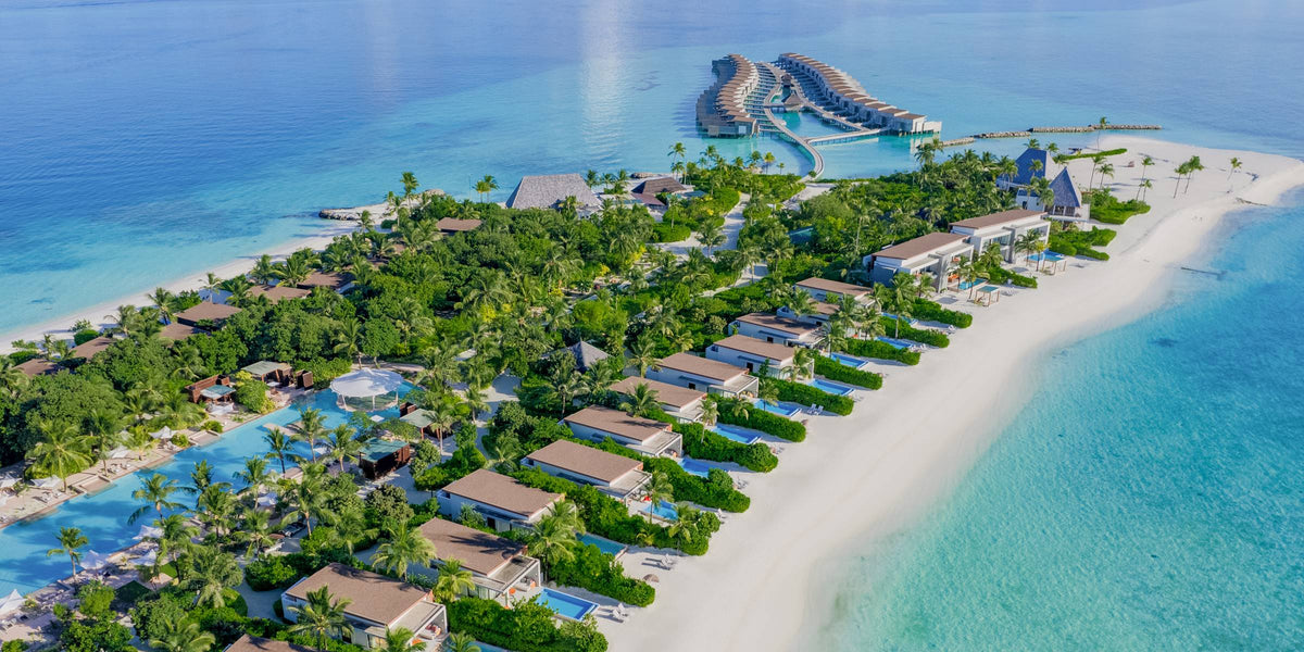 Kuda Villingili Resort from the air: beach villas with private pools set among palms along a wide white beach, with overwater villas in the lagoon beyond.