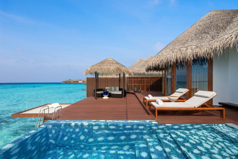The deck of an overwater villa at W Maldives, thatched roof above loungers and a timber sofa.