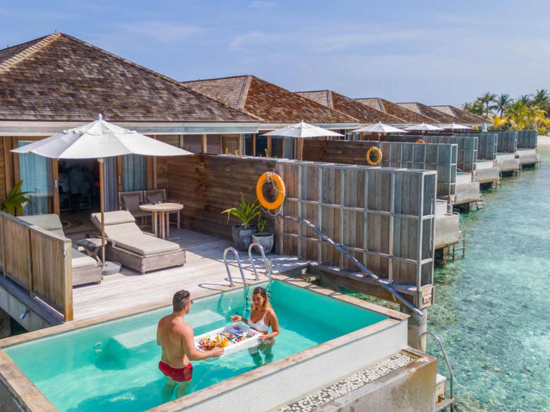 A couple sharing a floating breakfast tray in the private pool of an overwater villa at Kagi Maldives Resort & Spa, the row of villas stretching behind.