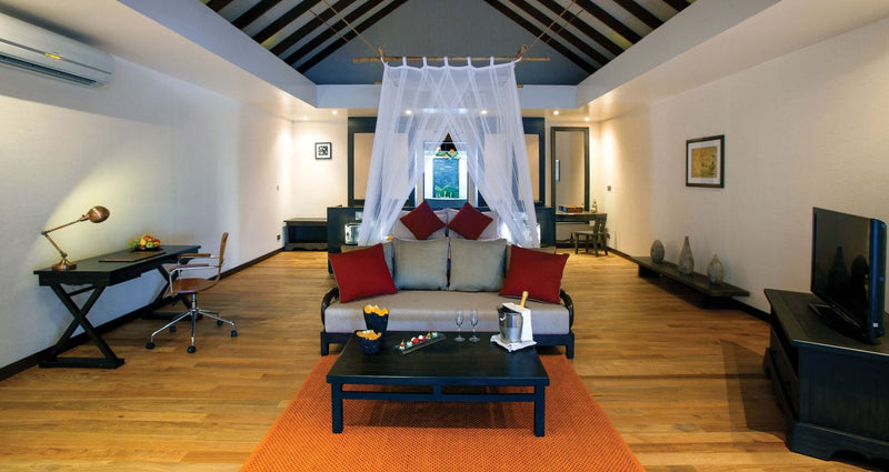 A beach villa interior at Atmosphere Kanifushi under a high pitched ceiling, a canopied bed with red cushions, a writing desk, a sofa and a low table on a wooden floor.