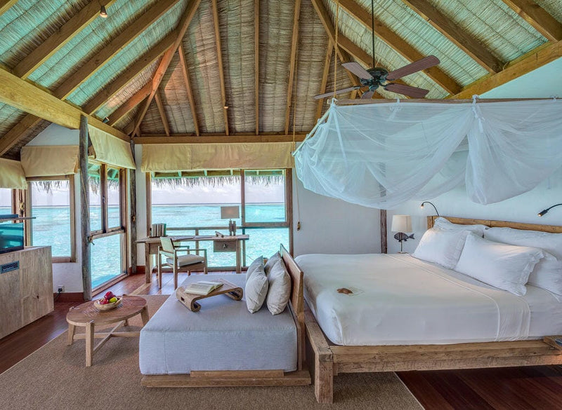 A bedroom at Gili Lankanfushi, four-poster bed draped in white netting under a pitched thatch roof.