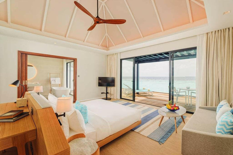 A water villa bedroom at Nova Maldives, sofa and low table, doors open to the deck and the lagoon.