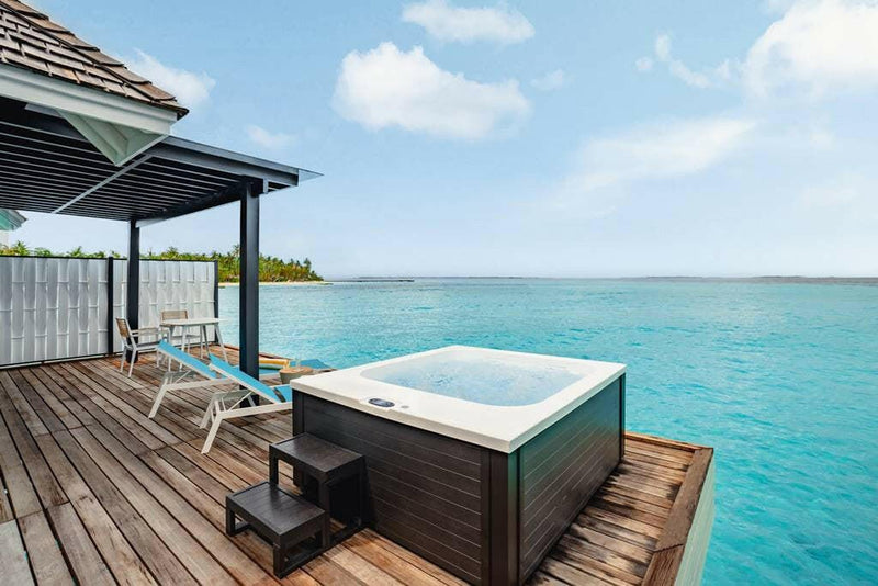 The deck of a water villa at Nova Maldives, a black jacuzzi tub and a lounger above open water.