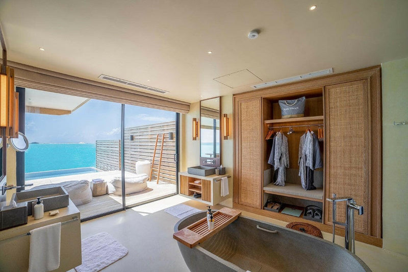 A dressing area at Kuda Villingili, robes in a timber wardrobe and a stone bath by the sea doors.