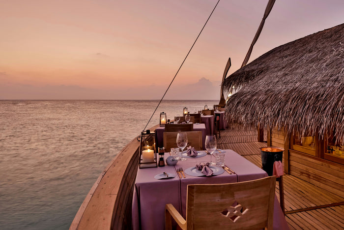 The deck of Ba'theli by the Reef at Milaidhoo Maldives at dusk, a candlelit table under the thatched roof beside the water