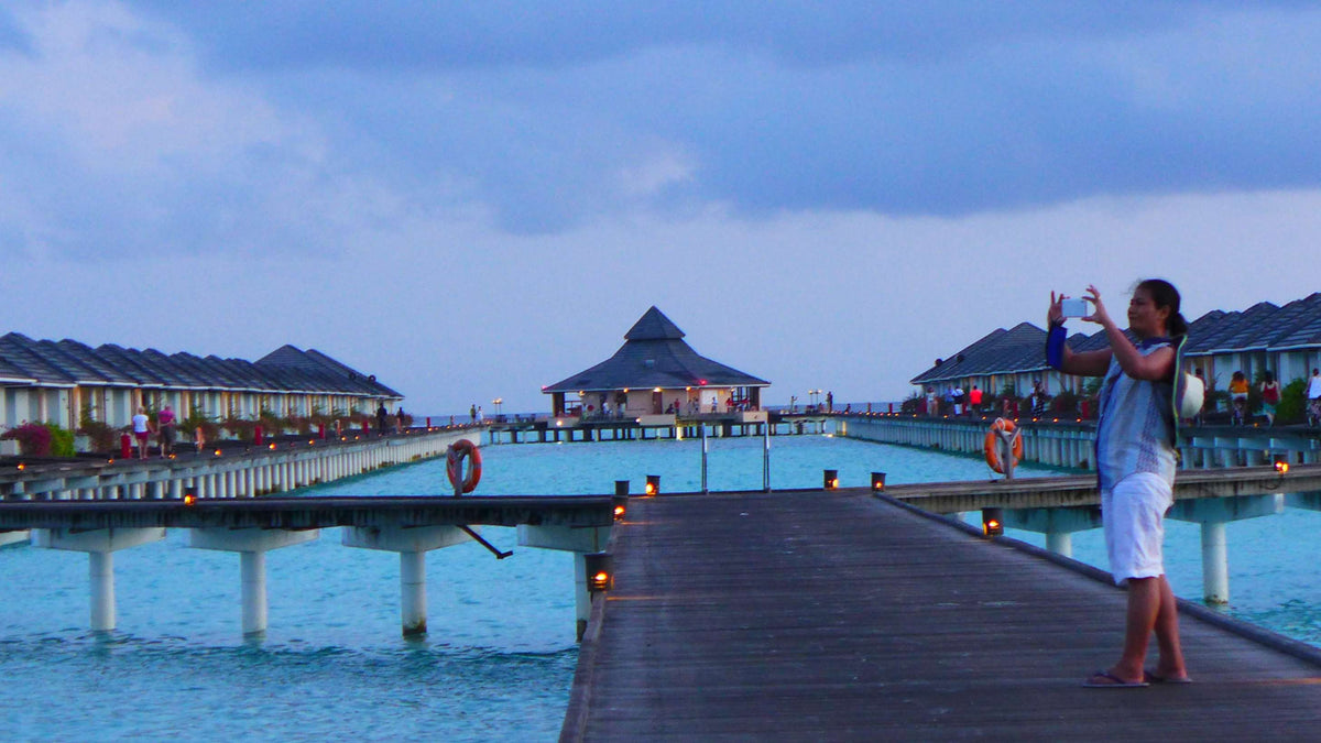 A woman photographing the water villas from a long jetty at Villa Park Sun Island at dusk, with lanterns along the walkway