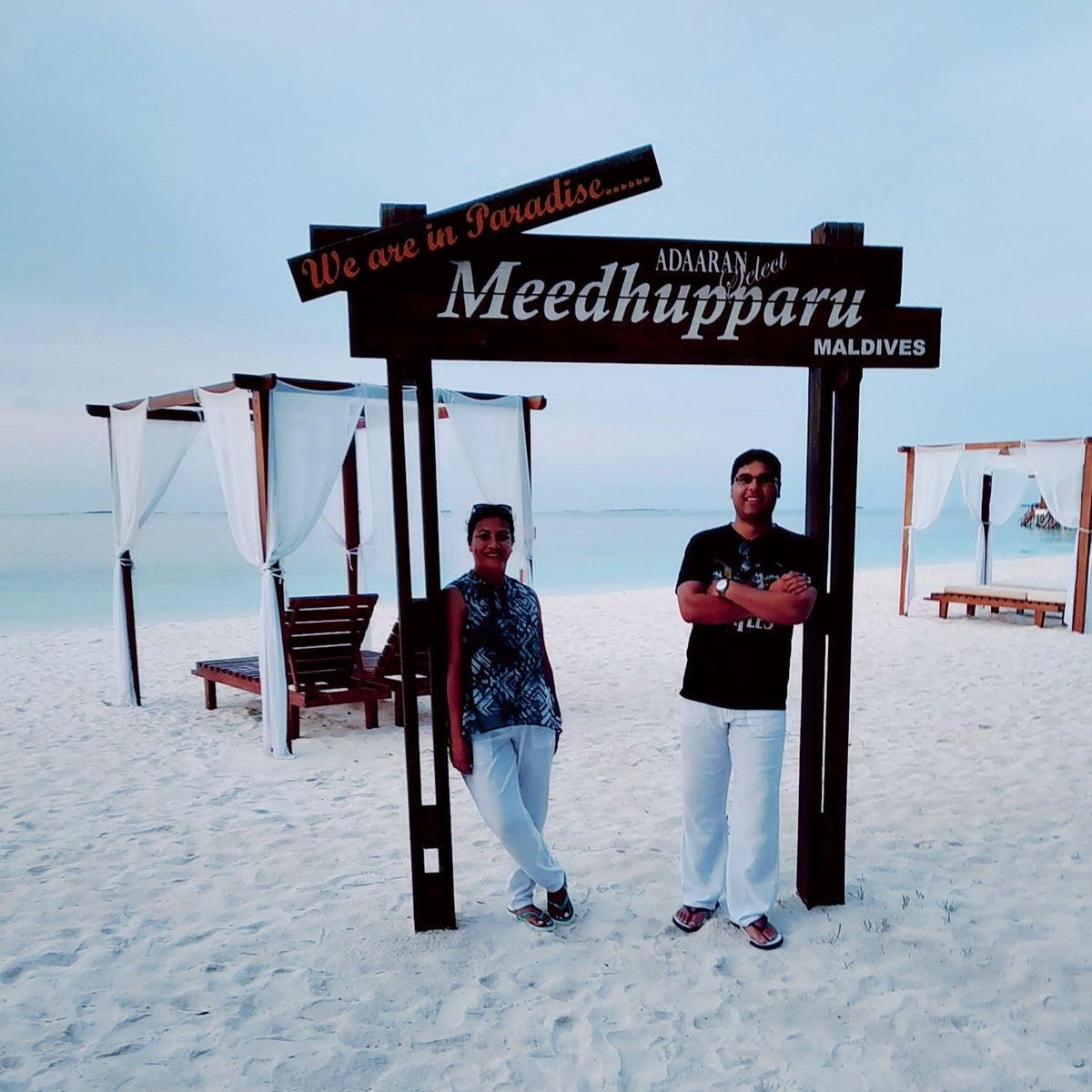 Two people on white sand under the Adaaran Select Meedhupparu sign, with white-curtained beach cabanas and a calm lagoon behind