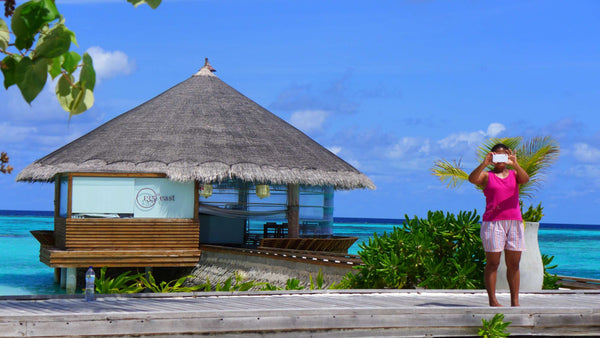 A woman photographing the view from a deck at Maafushivaru Maldives, beside a thatched overwater pavilion and a turquoise lagoon