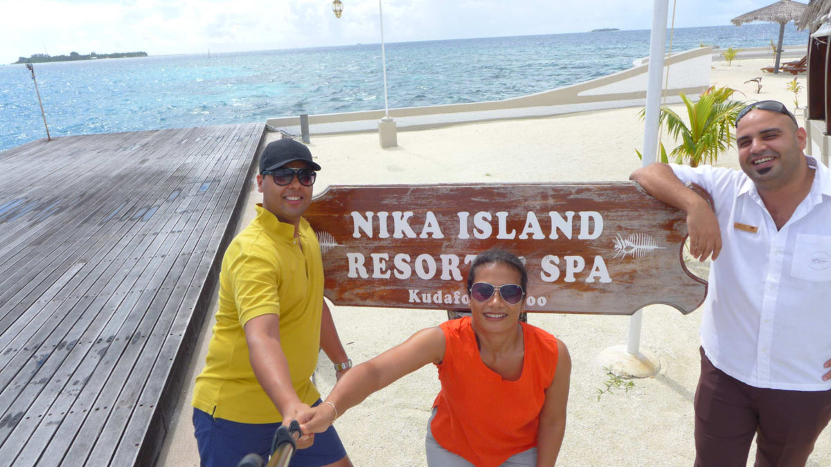 Three people, one of them resort staff, with the wooden Nika Island Resort & Spa sign on the beach