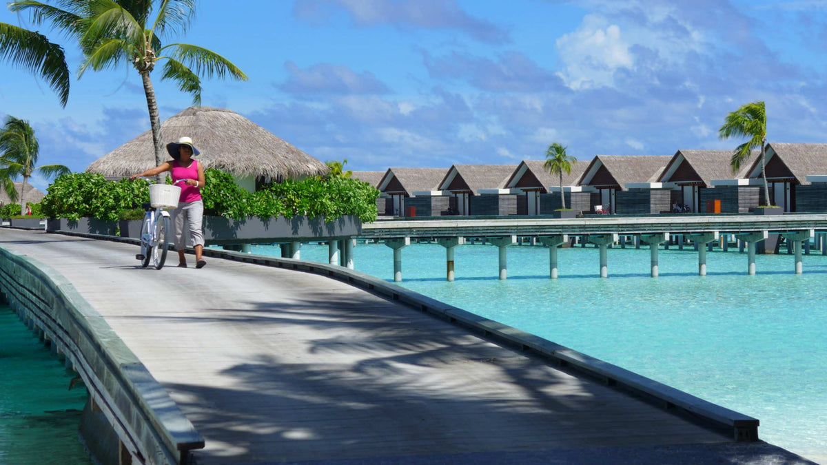 A woman with a bicycle on a boardwalk at Niyama Private Islands Maldives, with a thatched pavilion and water villas on a turquoise lagoon