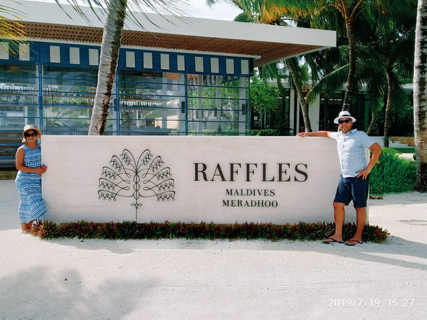 Two people either side of the Raffles Maldives Meradhoo sign, with a striped-awning building and palms behind