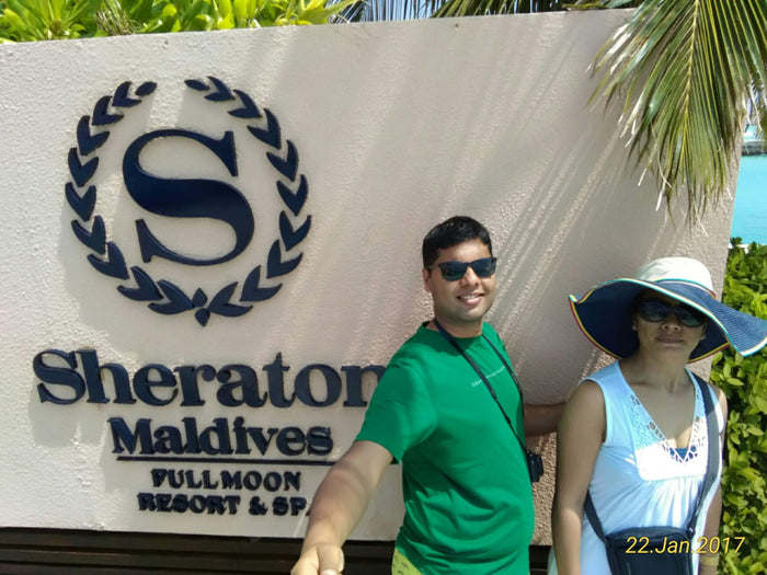 Two people in front of the Sheraton Maldives Full Moon Resort & Spa sign, beside palm leaves