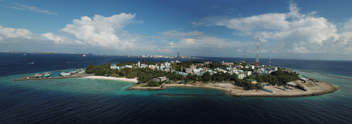 Villimalé from the air, a small green island ringed by beach and lagoon, with the Malé skyline on the horizon