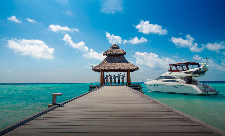 Staff waving from the thatched arrival pavilion at the end of the jetty at Baros Maldives, a motor yacht moored alongside in turquoise water.