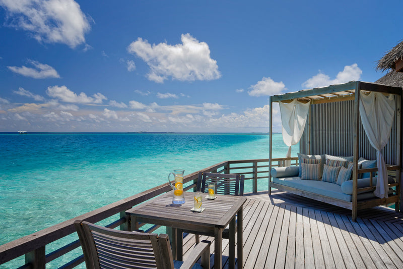 A Water Villa deck at Baros Maldives, a curtained daybed and a table for two above the turquoise lagoon.