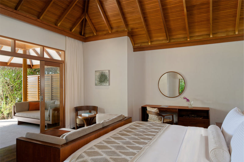 A Deluxe Beach Villa bedroom at Baros Maldives, a wide bed and sofa under a timber ceiling.