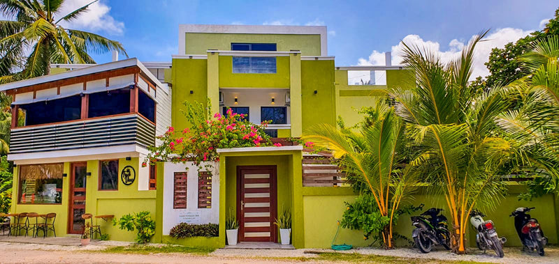 Front of Cozy Roots Fuvahmulah, a green and yellow building with a flowering garden