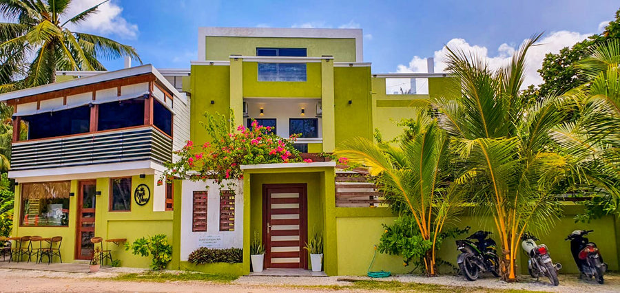 Front of Cozy Roots Fuvahmulah, a green and yellow building with a flowering garden