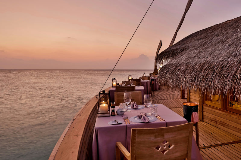 The deck of Ba'theli by the Reef at Milaidhoo Maldives at dusk, a candlelit table under the thatched roof beside the water