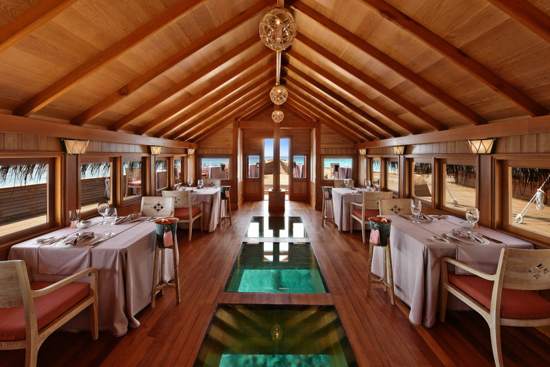 Ba'theli by the Reef at Milaidhoo Maldives, a timber-vaulted overwater dining room with a glass floor panel looking down onto the lagoon