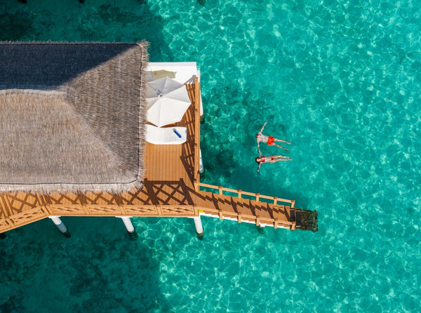 A premium water bungalow at Angaga seen from directly above, a swimmer floating in the turquoise beside its deck
