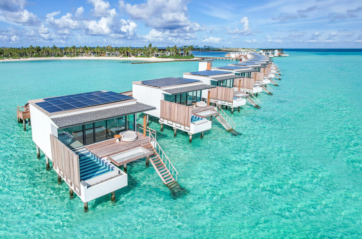 SO/ Maldives from the air, flat-roofed overwater villas with solar panels above a clear lagoon