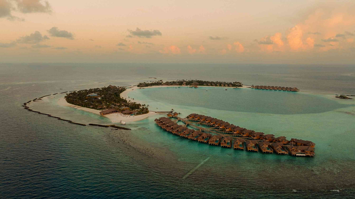ananea Madivaru Maldives from the air at sunset, the two islands joined by a sandbank with the overwater villas curving out into the lagoon