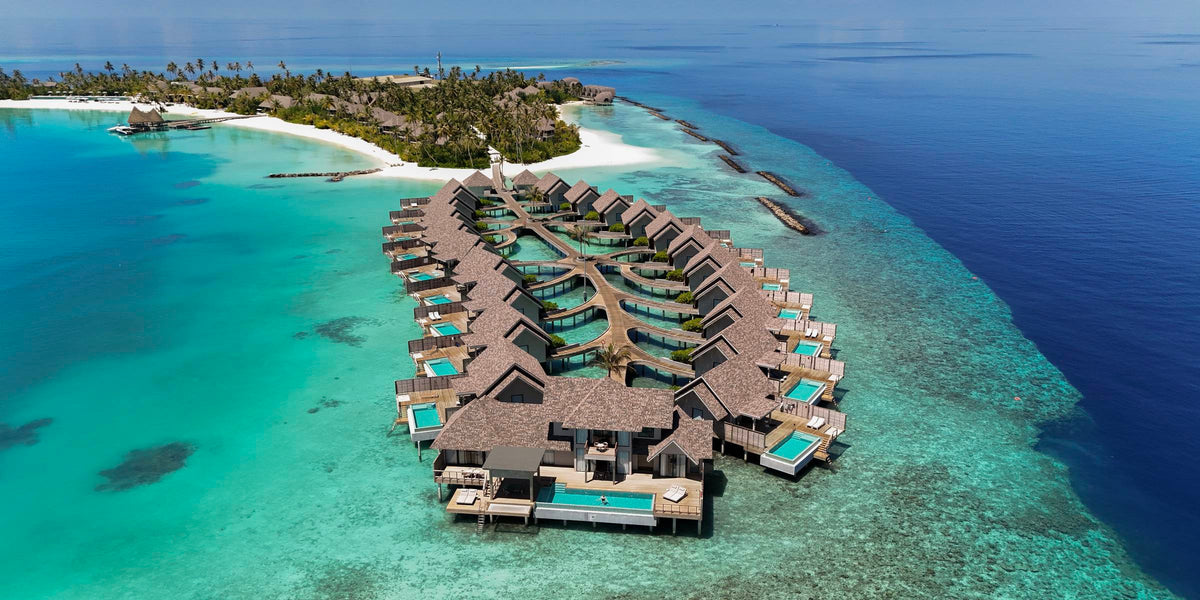 Aerial view of ananea Madivaru: two curved rows of thatched overwater villas, each with its own plunge pool, over a turquoise lagoon beside a palm-covered island