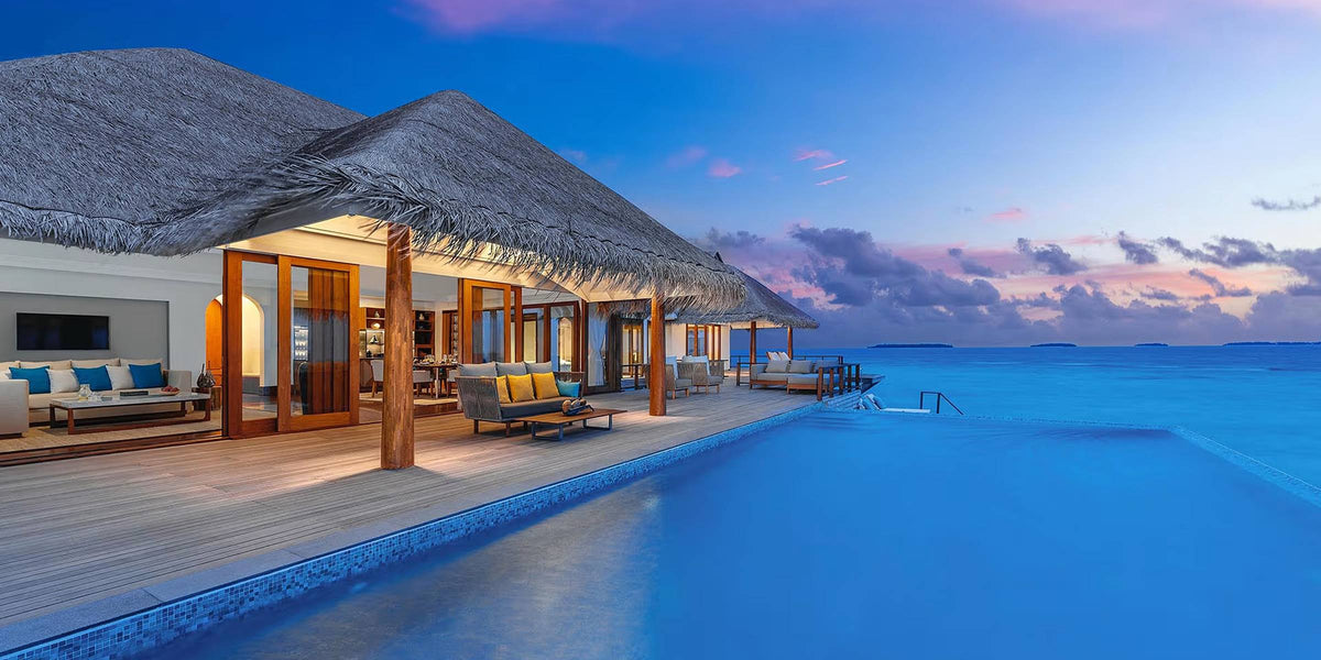 Anantara Kihavah Maldives Villas at dusk: a thatched overwater villa with lounge seating on a timber deck beside an infinity pool, the sea and a pink sunset sky beyond.