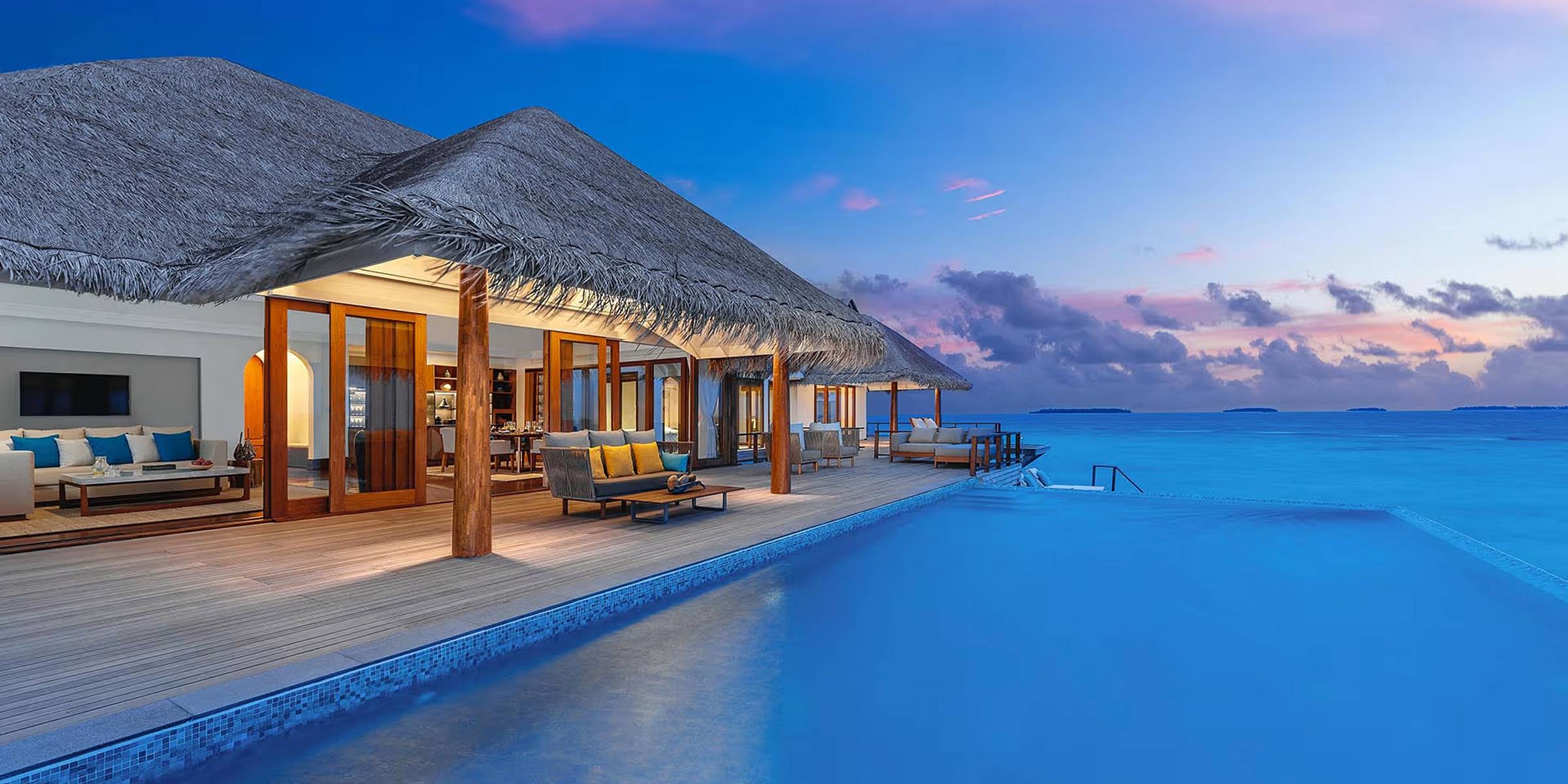 Anantara Kihavah Maldives Villas at dusk: a thatched overwater villa with lounge seating on a timber deck beside an infinity pool, the sea and a pink sunset sky beyond.