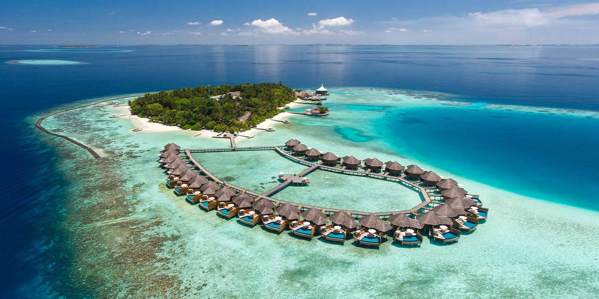 Baros Maldives from the air: a small wooded island with a loop of thatched overwater villas on the reef, the pale lagoon and the reef edge clearly visible.