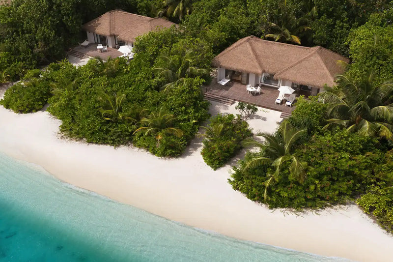 Beach villas at Noku Maldives from the air, thatched roofs in dense palms above white sand.