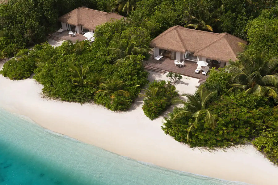Beach villas at Noku Maldives from the air, thatched roofs in dense palms above white sand.