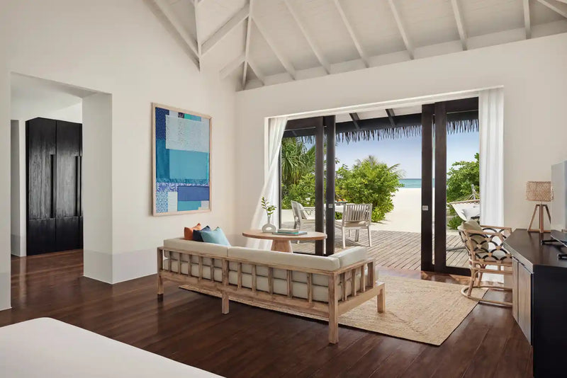 A beach villa interior at Noku Maldives, doors open onto a timber deck and the white beach.