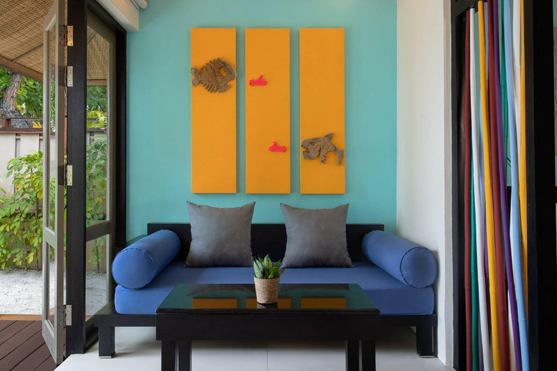 A villa daybed at Dhawa Ihuru, grey cushions below an orange fish triptych on a turquoise wall.