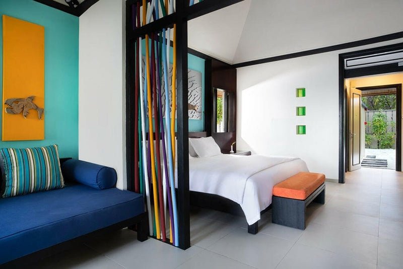 A villa interior at Dhawa Ihuru, blue daybed and striped curtain beside a bed with an orange bench.