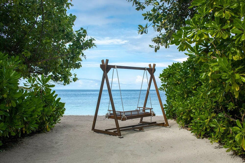 A wooden swing on the sand at Dhawa Ihuru, framed by green bushes with calm sea beyond.