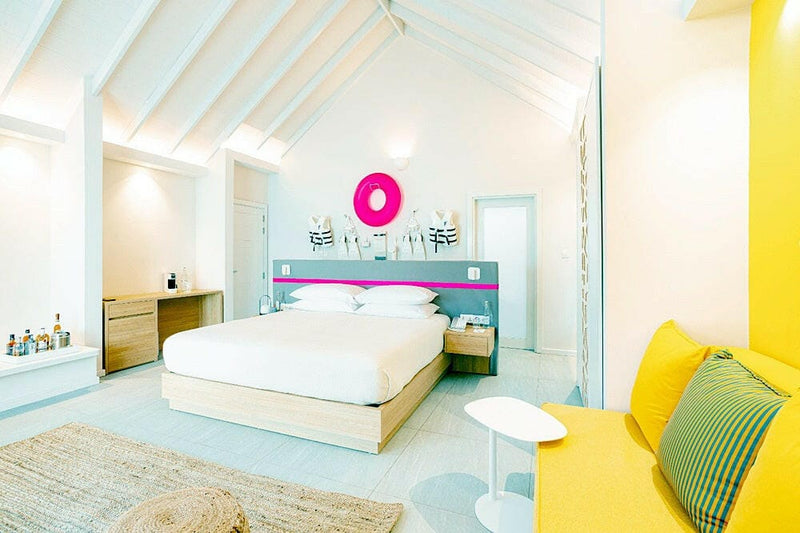 A villa bedroom at The Standard Huruvalhi, a pink neon ring above the bed and a yellow banquette alongside.
