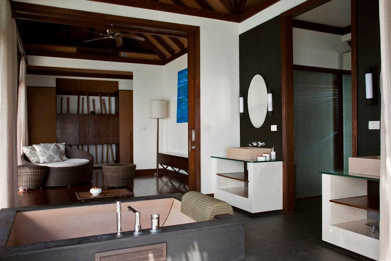 A villa bathtub at Coco Bodu Hithi below an oval mirror, the sitting room and its rattan daybed beyond