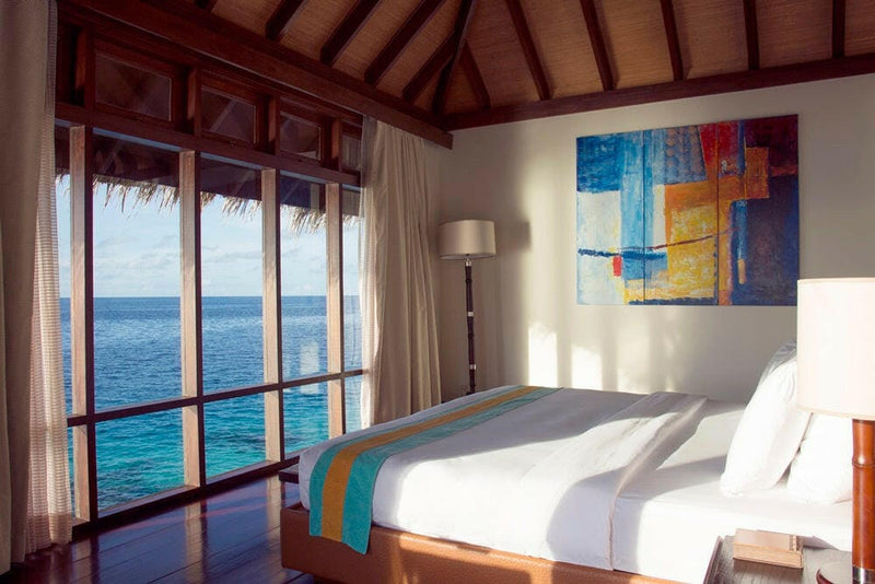 A water villa bedroom at Coco Bodu Hithi, a bright abstract canvas above the bed and glass doors to the lagoon
