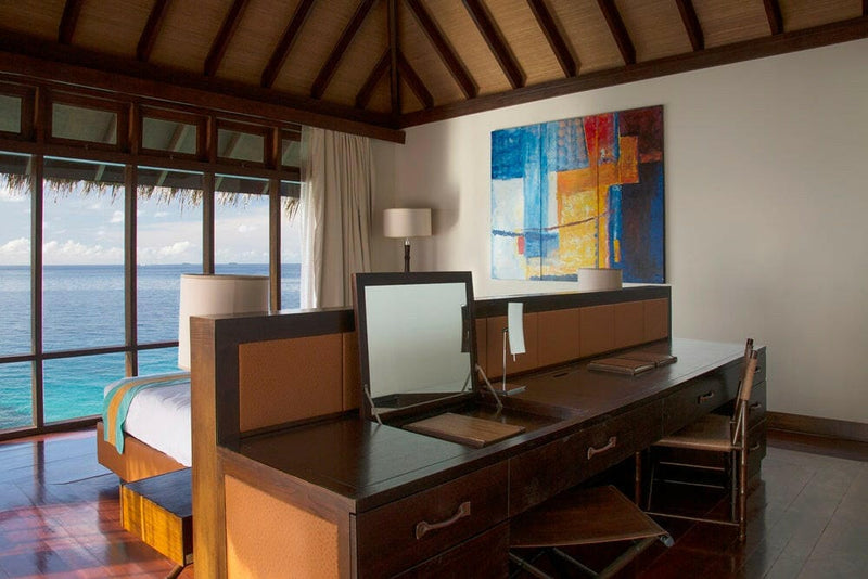 A writing desk beside the bed in a water villa at Coco Bodu Hithi, the lagoon through full-height glass