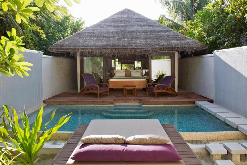 A walled garden pool at Coco Bodu Hithi, with a thatched pavilion and a pink cushioned daybed beside it