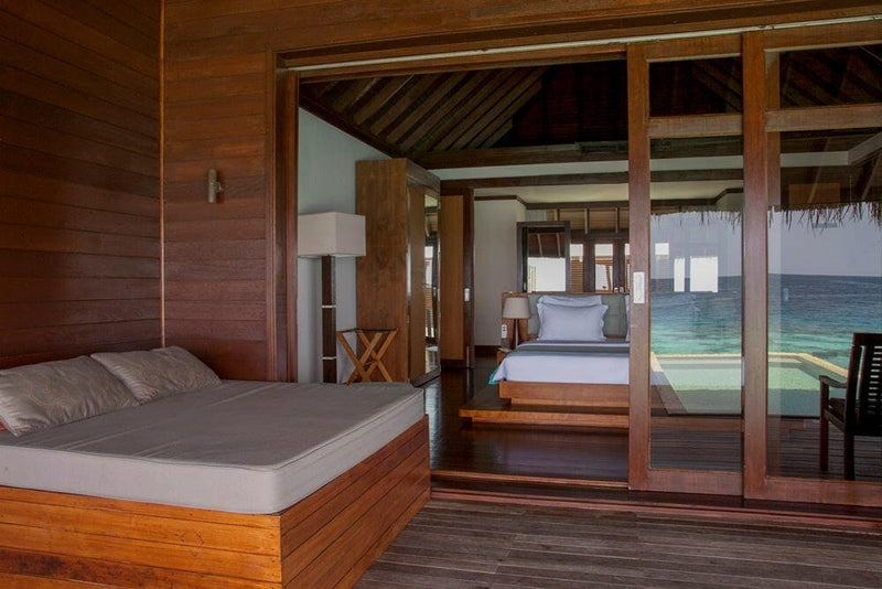 A water villa bedroom at Coco Bodu Hithi, timber-lined walls and sliding doors open onto the deck