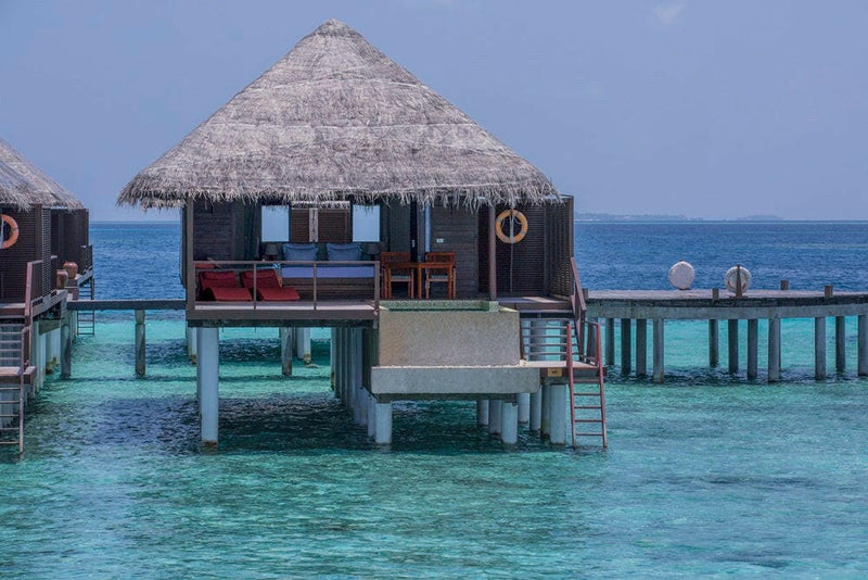 A water villa at Coco Bodu Hithi seen from the lagoon, red loungers on its deck under a thatched roof