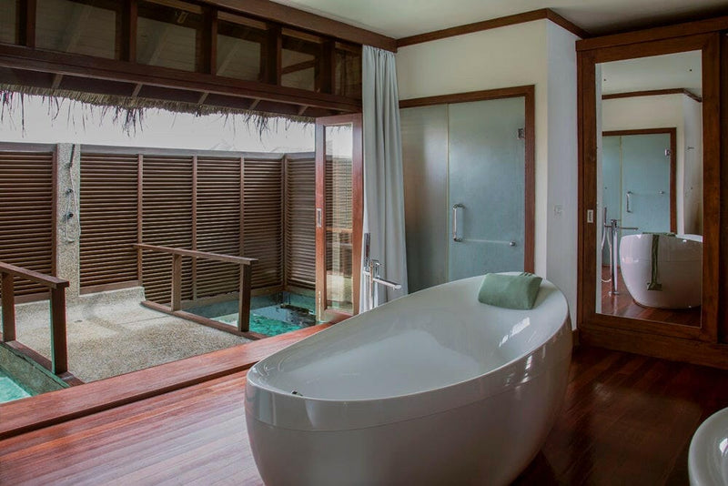 A white oval tub in a water villa bathroom at Coco Bodu Hithi, louvred screens opening to the deck
