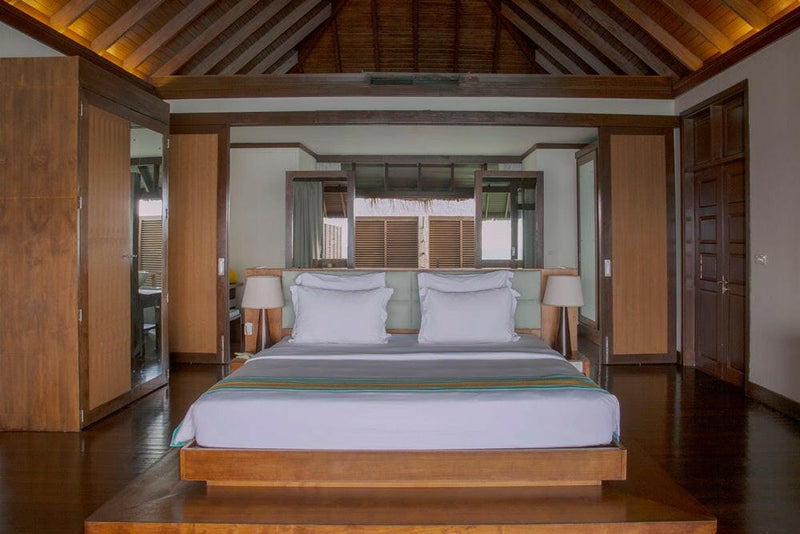 A water villa bedroom at Coco Bodu Hithi, a king bed in white linen between two timber-lined walls