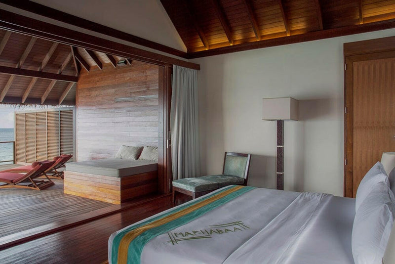 A water villa bedroom at Coco Bodu Hithi, a welcome runner on the bed and louvred doors open to red loungers