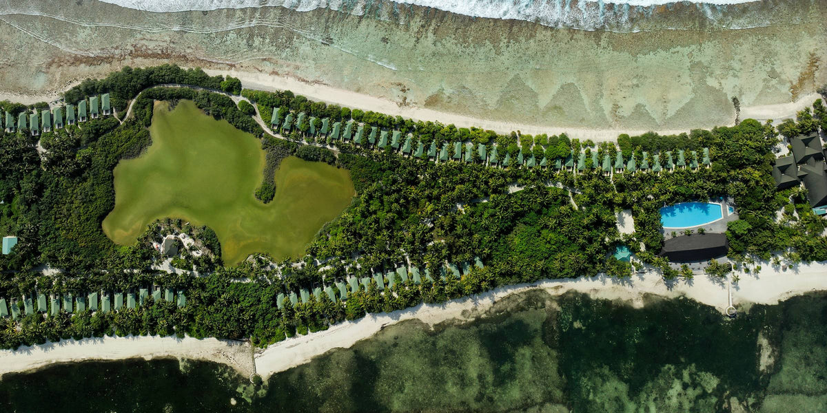 Canareef Resort from directly overhead: a long narrow island of dense palm forest with a green inland lagoon at its centre, villas lining both shores, and reef showing dark through the water off the beach.