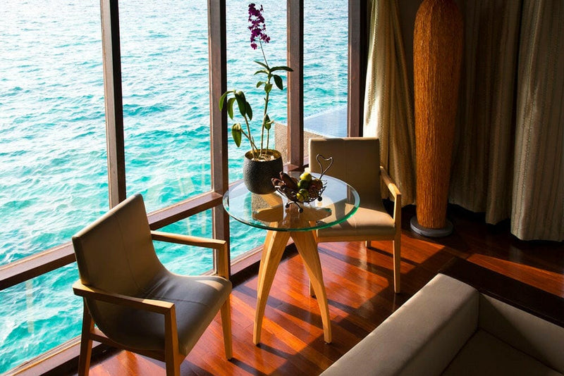 Two timber armchairs and an orchid on the table in a villa at Coco Bodu Hithi, lagoon through the glass