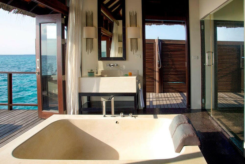 A villa bathroom at Coco Bodu Hithi, a wide cream tub in front of twin basins and doors onto the deck